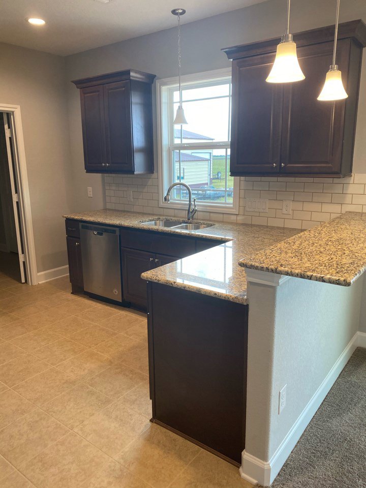 Kitchen with dark wood cabinets, granite countertops, stainless steel sink, pendant lights, white walls, and a window providing natural light