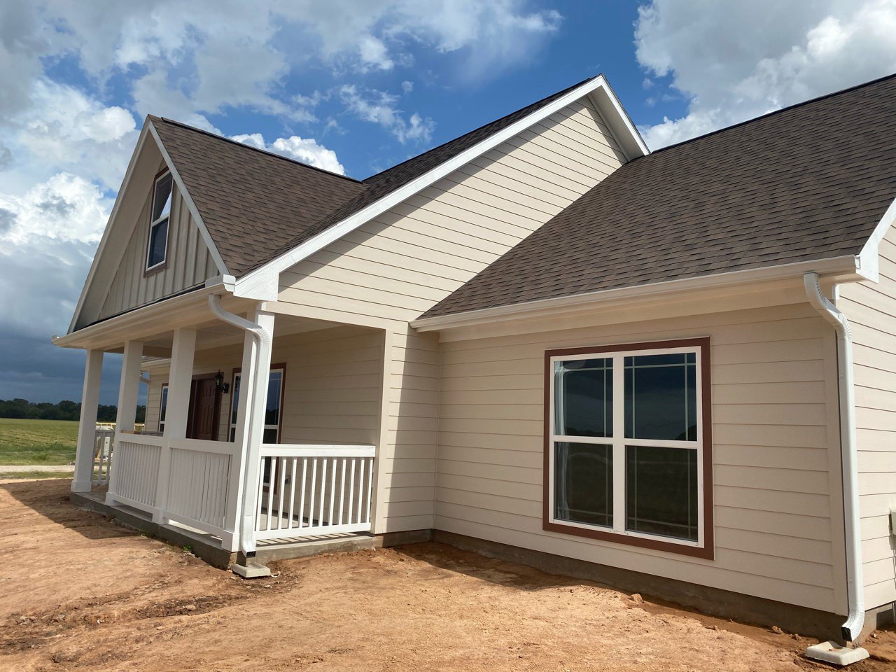 Partially built house with white porch railing, white-trimmed window, white door, and exposed dirt yard under cloudy sky