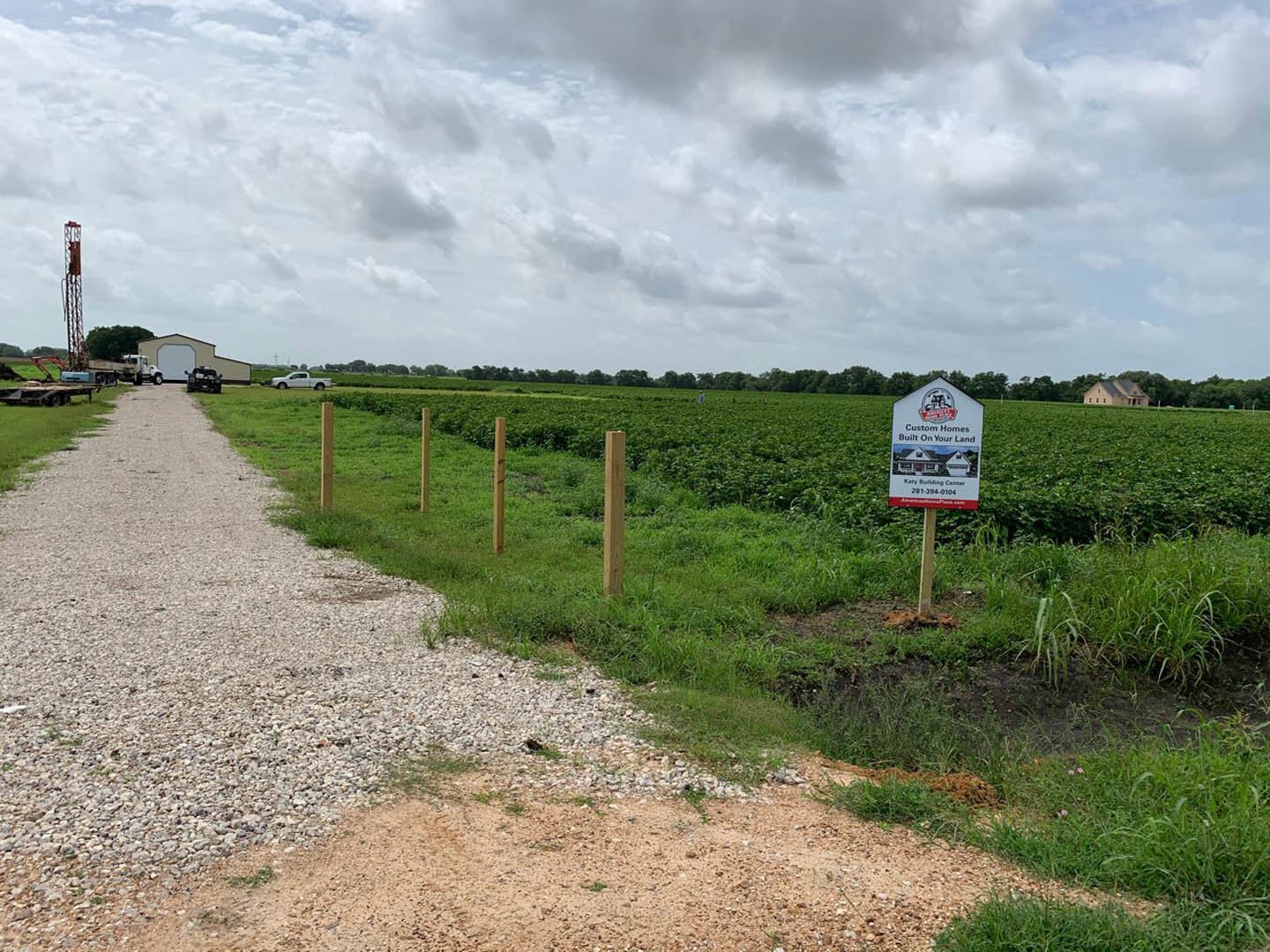 White sign with house illustration mounted on wooden post in grassy field, gravel driveway leading to garage with parked truck, row of cars in background, cloudy sky overhead