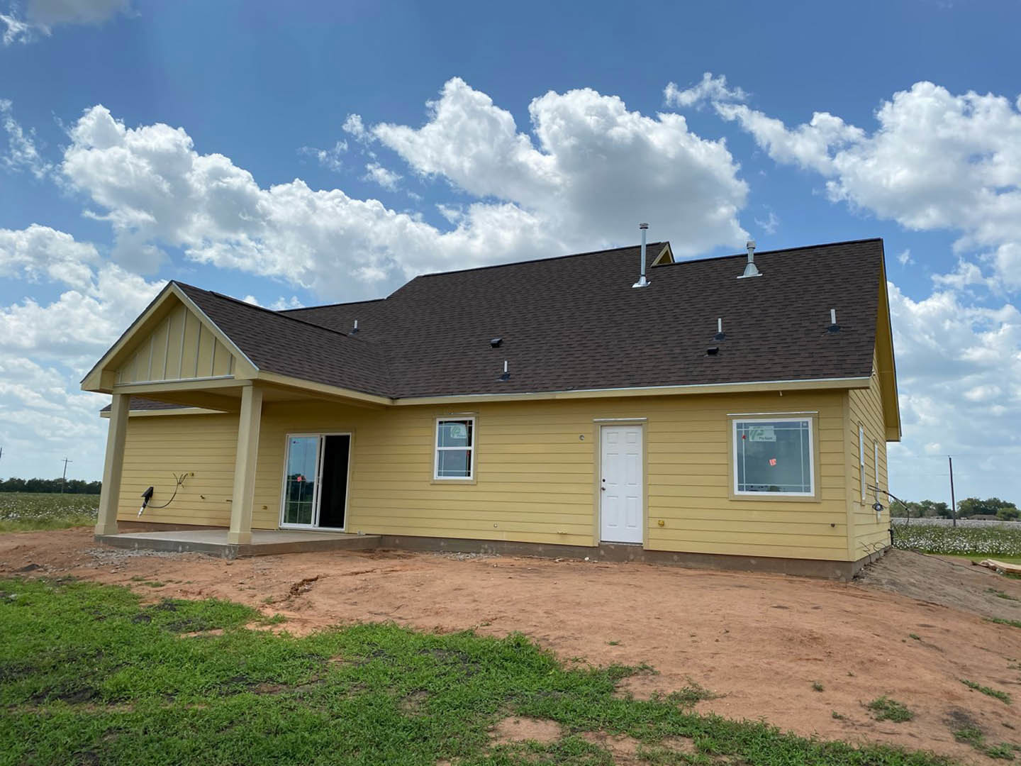 Yellow siding house with white door, black handle, and single window displaying a sticker, set against blue sky and dirt yard.