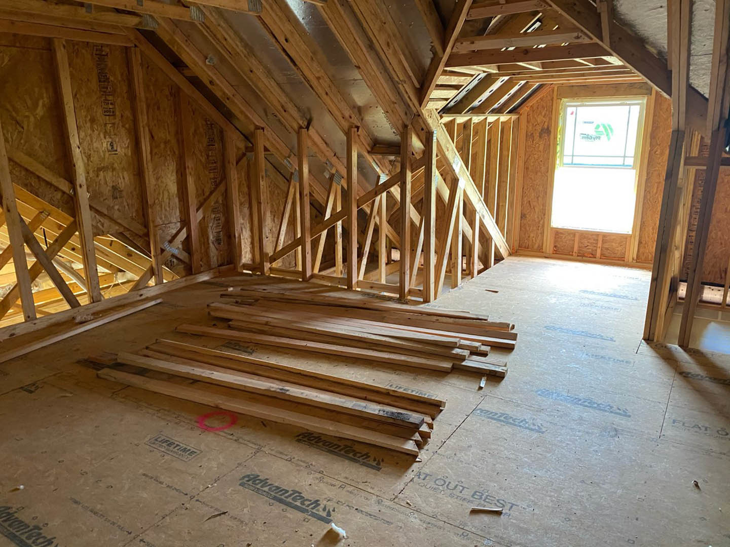 Exposed wooden beams across ceiling, hardwood planks stacked on floor, close-up of window with logo, unfinished attic interior with building insulation visible