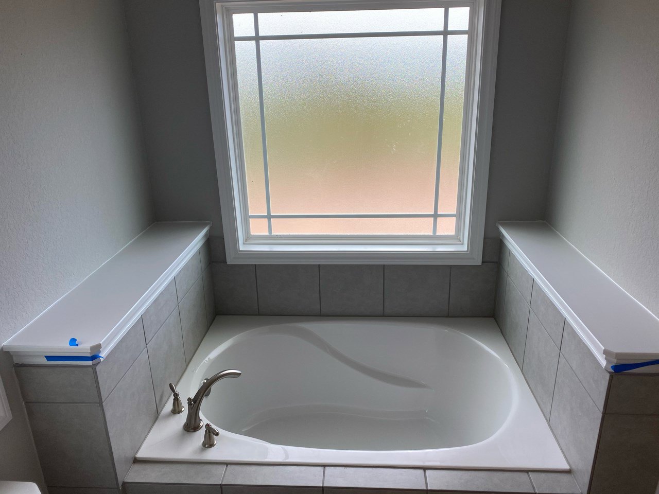White freestanding bathtub on gray tile floor beneath frosted glass window, chrome faucet, white shelf with blue trim along wall