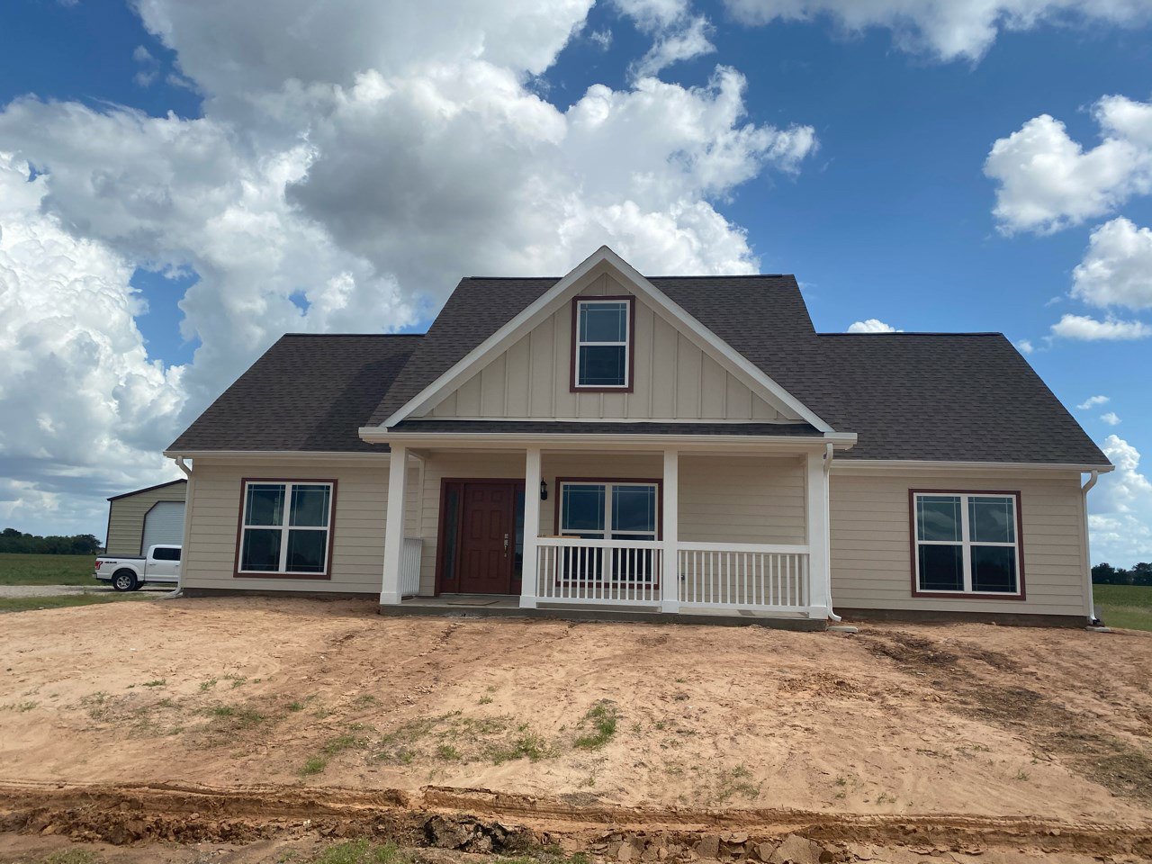 Two-story house under construction with exposed siding, covered porch, white-framed window, brown door with glass pane, dirt patch in front, white truck parked nearby, cloudy sky