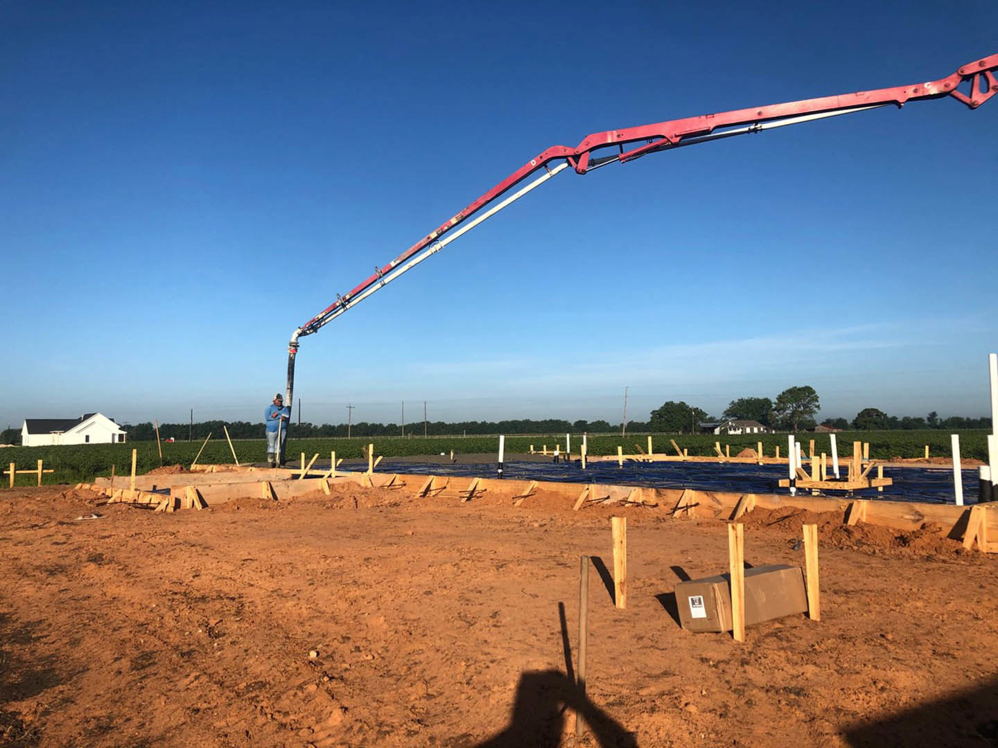 Red crane positioned above a dirt construction site with wooden posts, white house featuring black roof in background, man operating crane under clear blue sky.