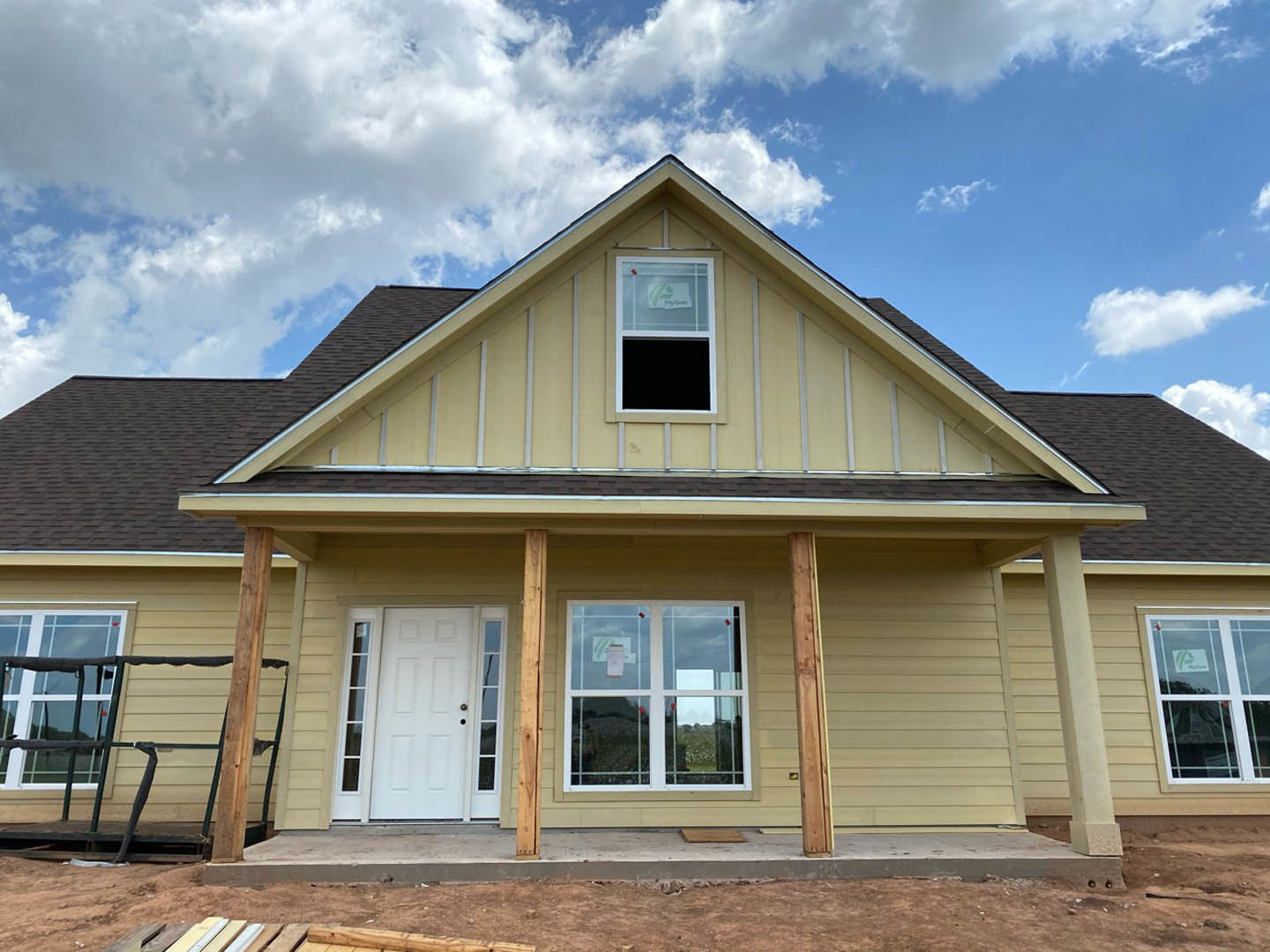 Two-story house under construction with light-colored siding, white door with glass panels, multiple windows including one with a sign, unfinished railing, and blue sky overhead