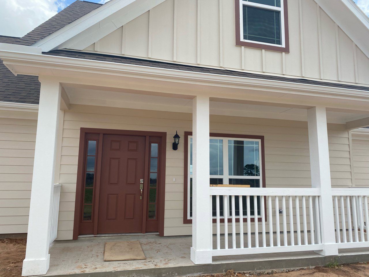 Front porch with white railing, brown wooden door featuring glass panels, white-framed windows, light siding, and a tree visible outside