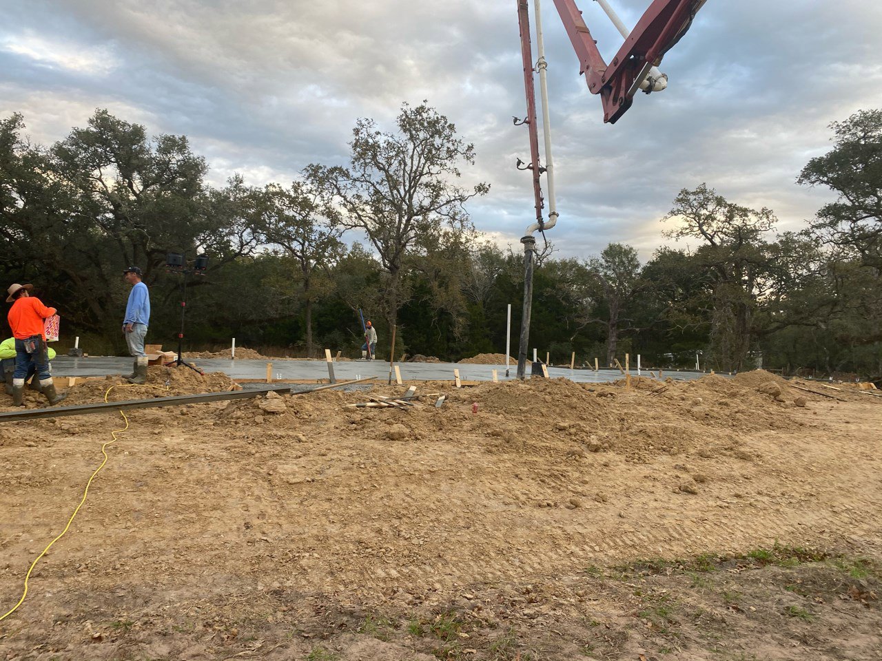 Dirt construction site with crane, scattered poles, yellow cable, and workers in orange and blue shirts; surrounded by trees under cloudy sky