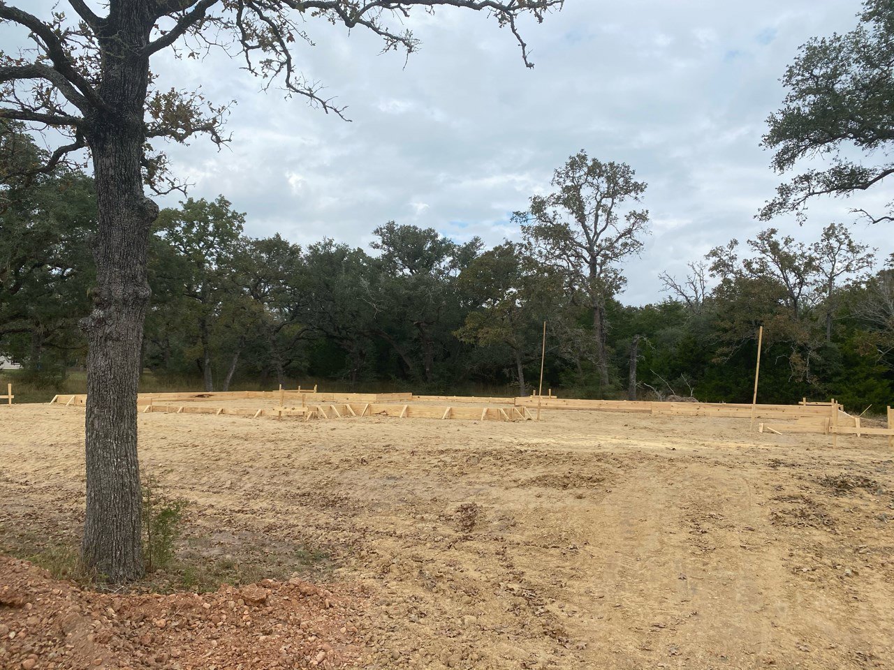 Wooden framing on a dirt lot surrounded by grassy field, scattered trees, and cloudy sky