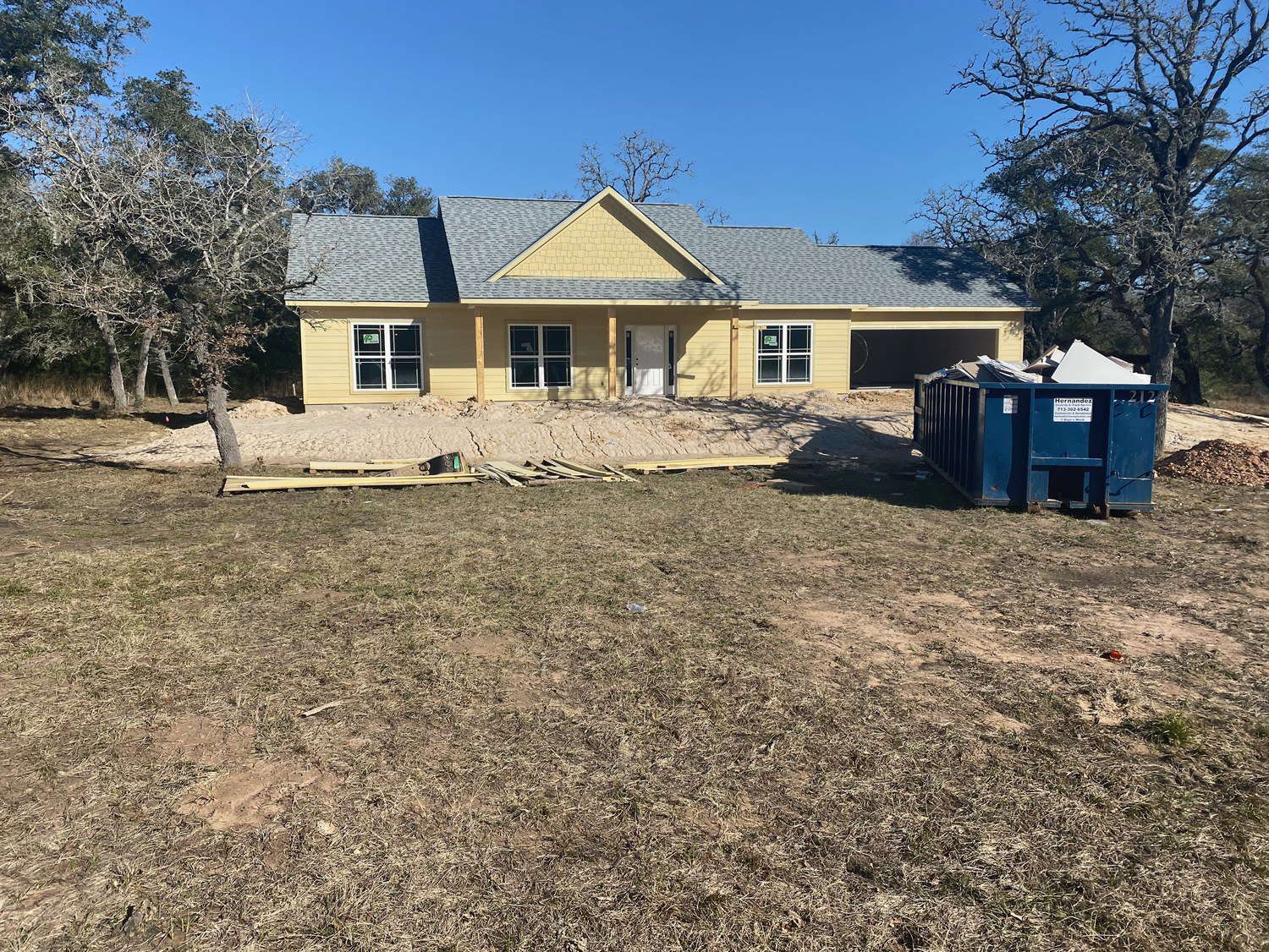 White house with black trim and white door, dry grass yard scattered with wood debris, blue dumpster with sign, window displaying notice, clear blue sky overhead.