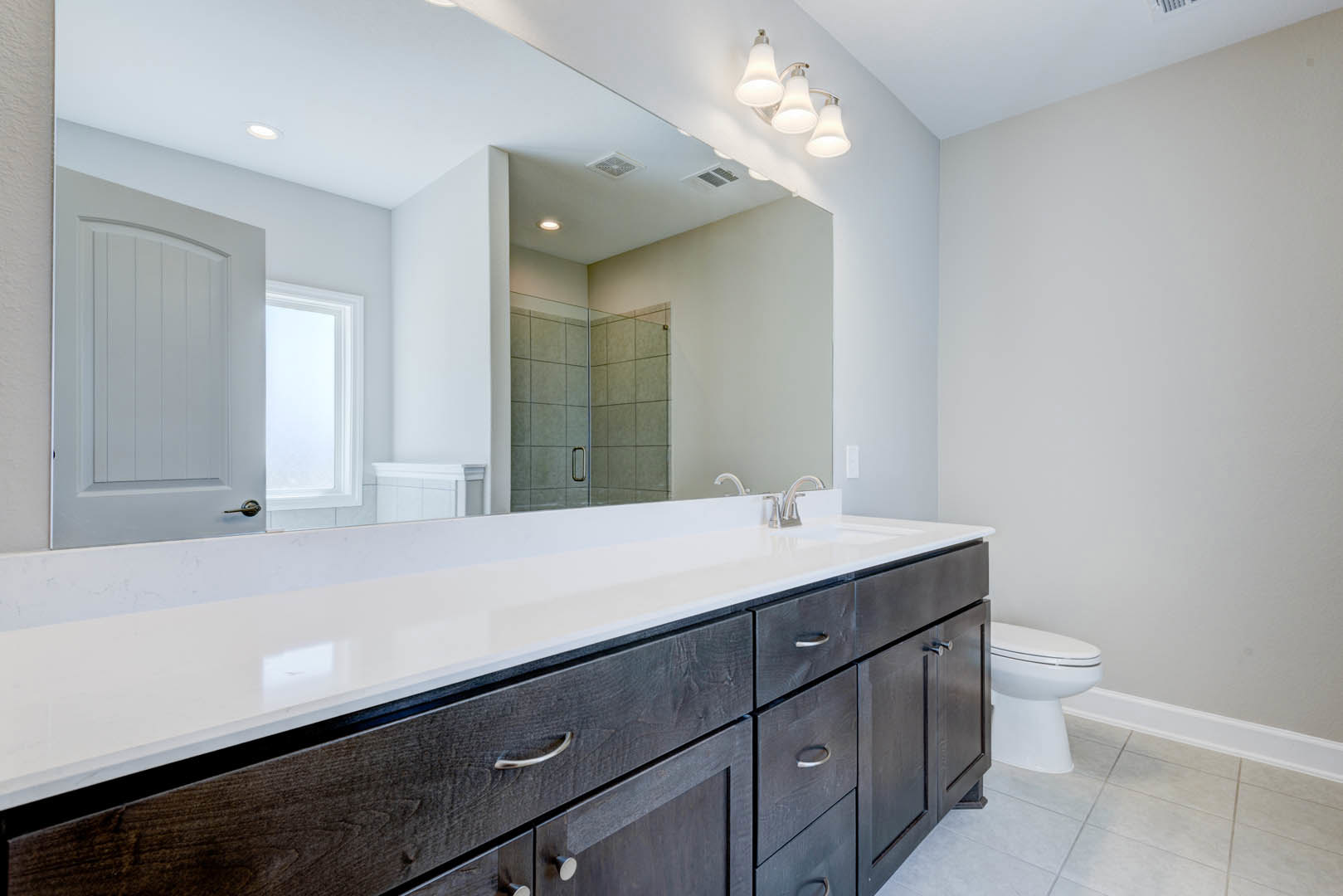 Bathroom with a wide mirror above a stone countertop, white toilet with closed seat, tiled walls, cabinetry with drawers, arched white door, window with trim, and modern light