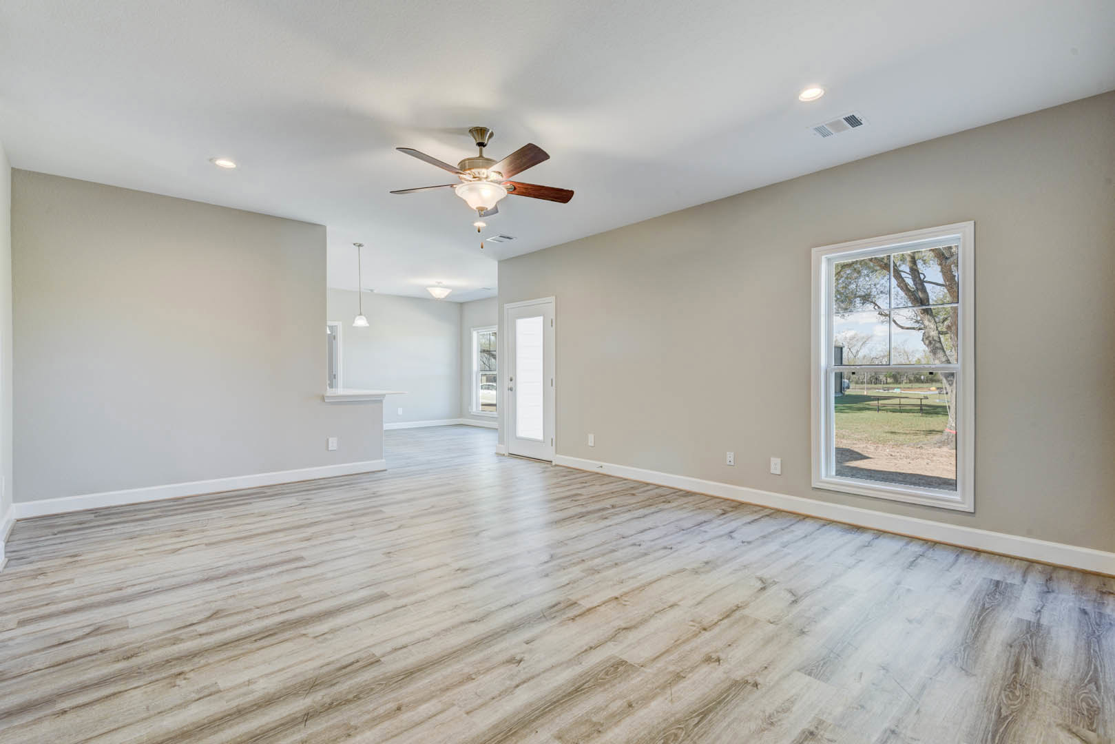 Hardwood floor room with ceiling fan, white walls, glass-paneled door, large window overlooking park and tree