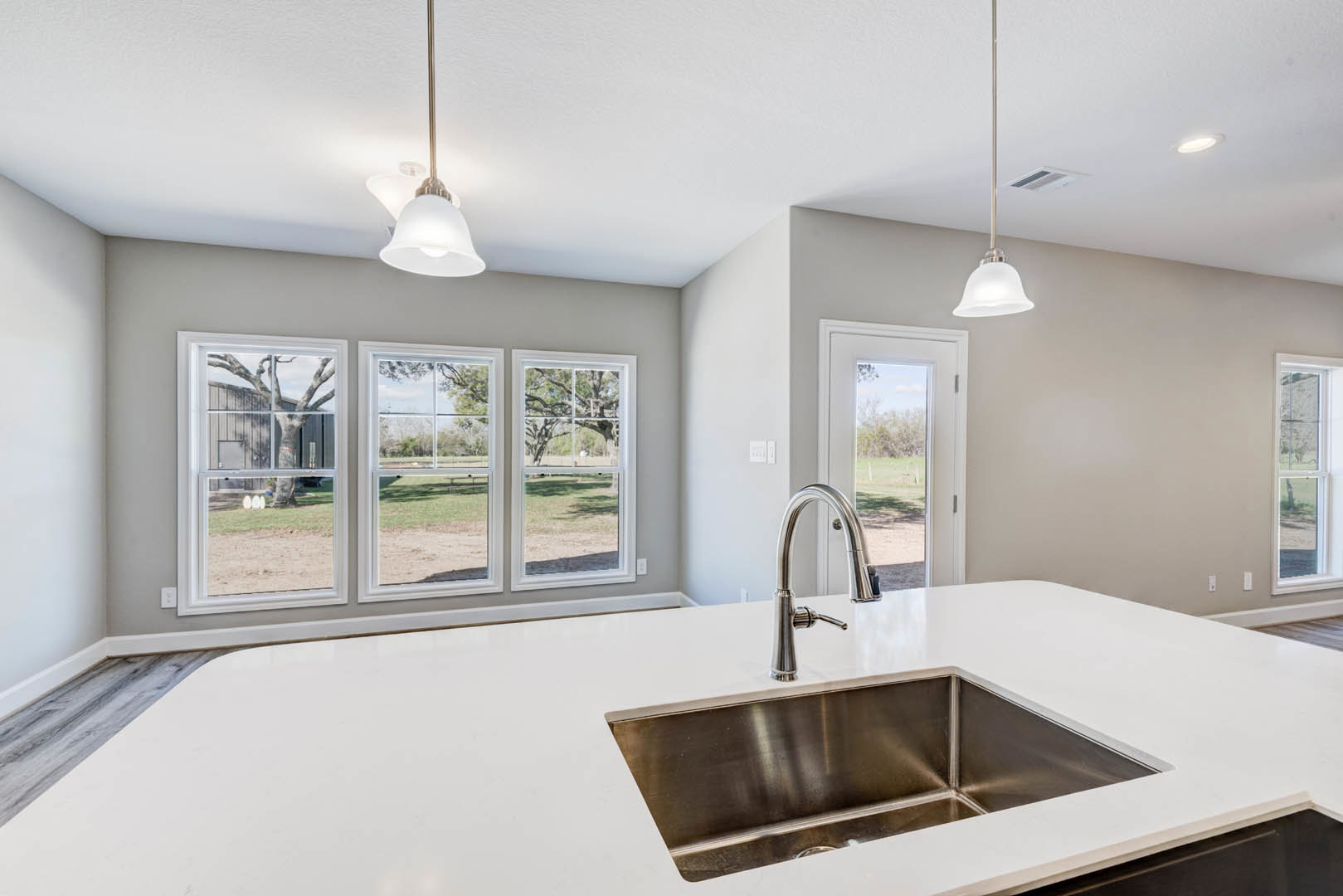 Stainless steel sink with modern faucet set in a light-colored countertop, row of large windows overlooking a green field, white cabinetry and minimalist light fixture visible