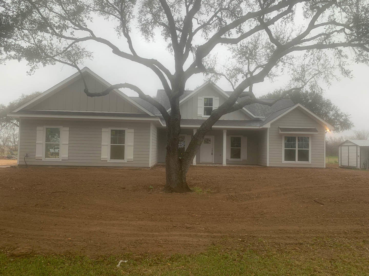 Two-story farmhouse with white siding, large front window with white frame, small shed to the side, mature tree planted in dirt near grassy lawn, blue sky overhead