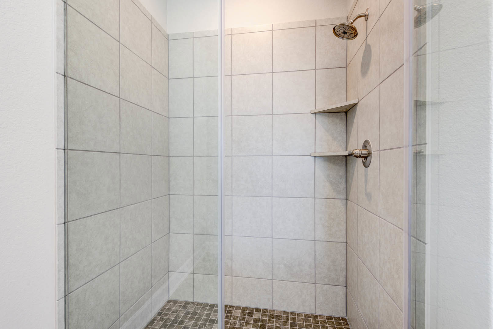 Glass shower enclosure with chrome shower head, brown and white tile floor, marble tile walls, built-in shelf, and exposed plumbing pipe.