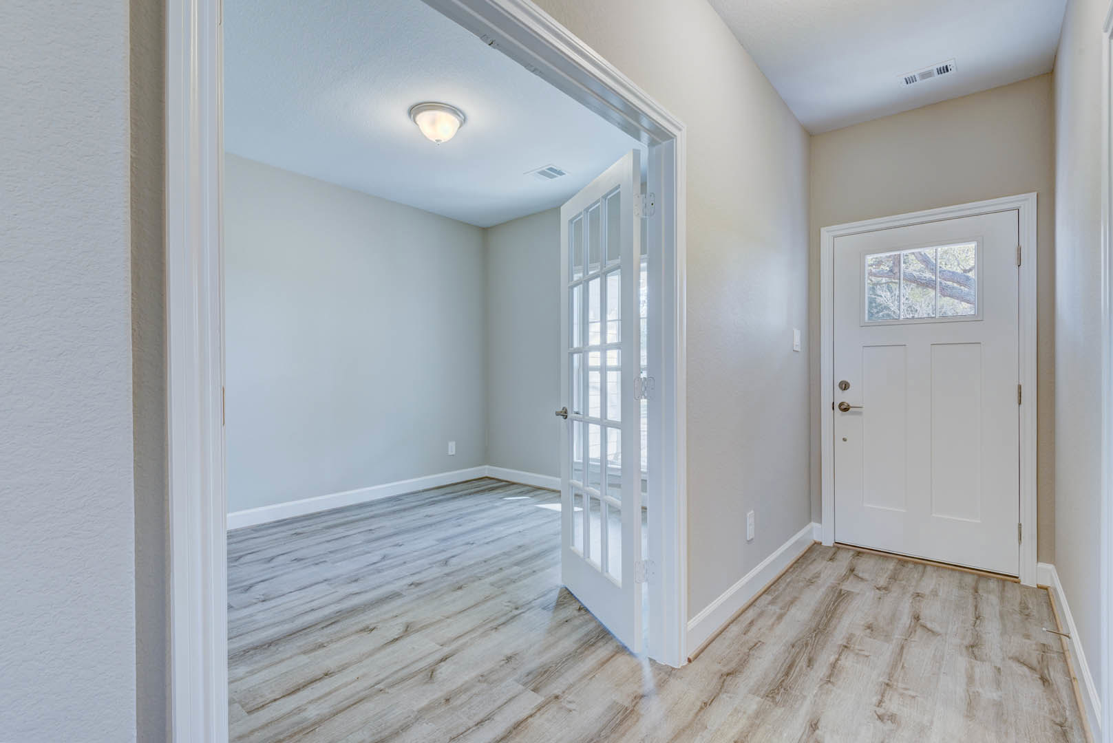 White paneled door with glass panes, wood laminate flooring, white plaster walls, ceiling light fixture, window with tree branch visible outside