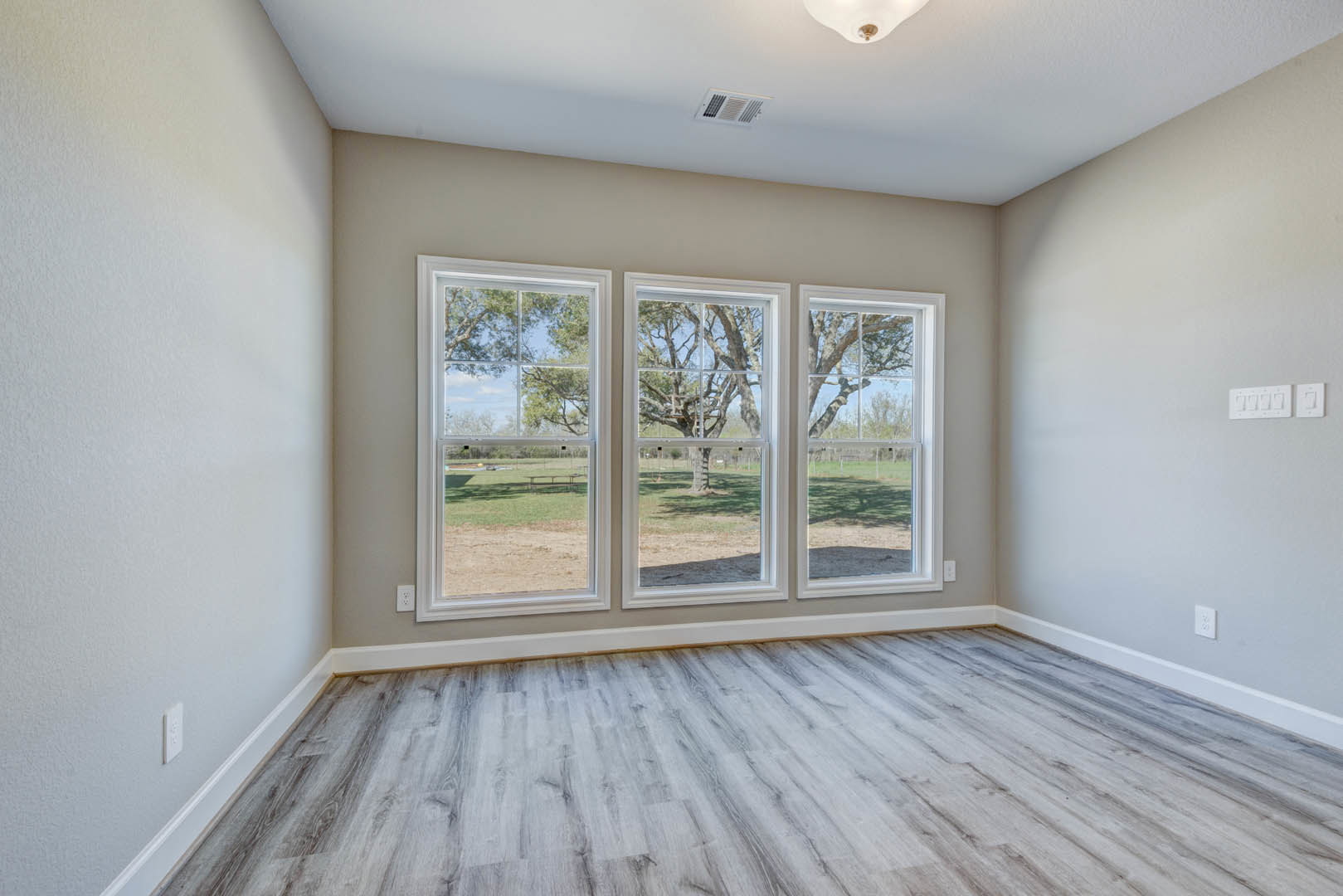 Sunlit room with wide wood plank flooring, large windows overlooking green grass and trees, white plaster walls, and a white light switch.