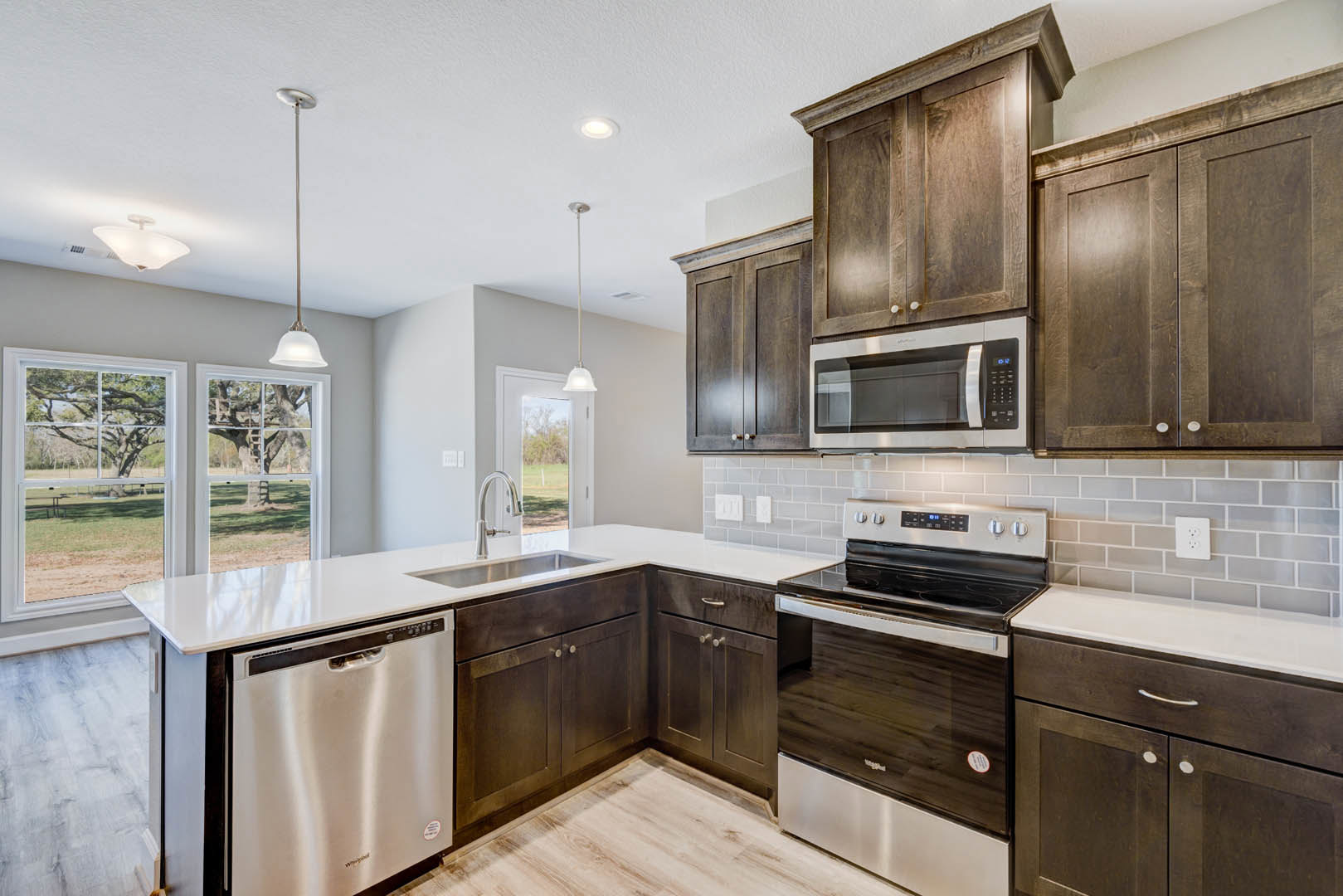 Kitchen with dark wood cabinetry, stainless steel appliances including microwave and stove, white wall outlet, window overlooking trees, and a tall flagpole visible outside