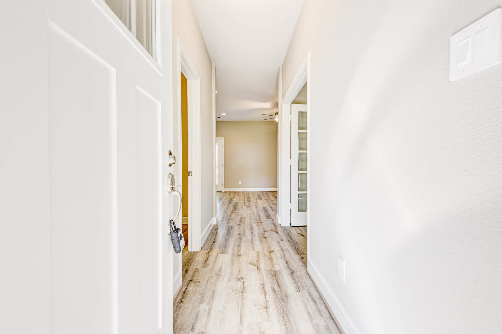 Hallway with smooth white walls, natural wood flooring, white door with keyhole, and modern light switch