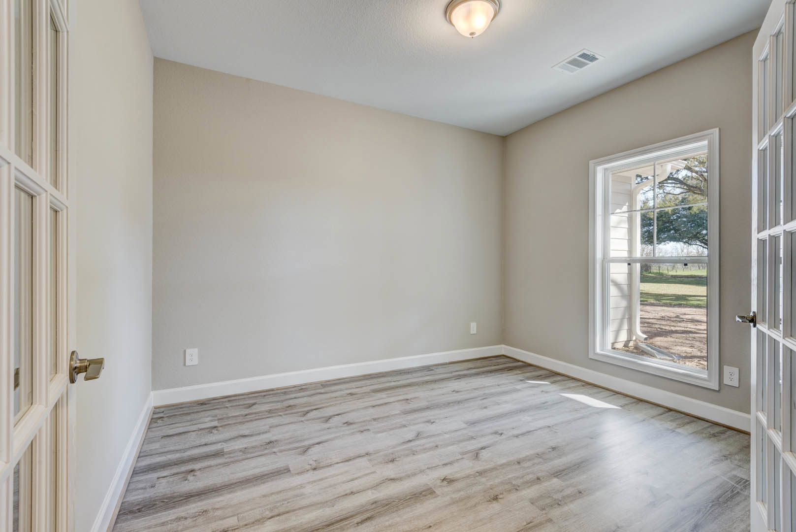 Sunlit room featuring wide wood plank flooring, crisp white walls, large window overlooking grassy field, and decorative white column with crown molding.
