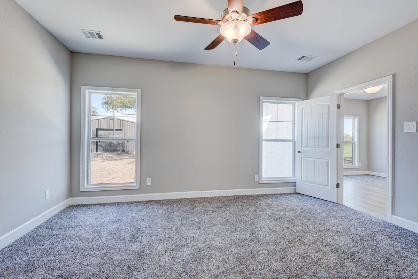 Neutral-toned room with carpet flooring, ceiling fan with light fixture, white door, window showing exterior building, and light switch on the wall