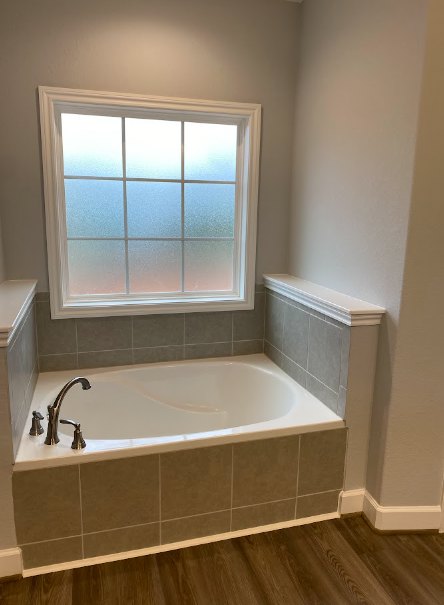 Freestanding bathtub with chrome faucet, wood floor, white tile walls, frosted glass window, and built-in white shelf