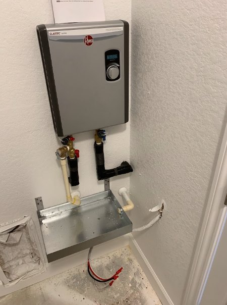 Water heater with exposed pipes beside a metal utility sink and a grey rectangular control unit in a tiled room