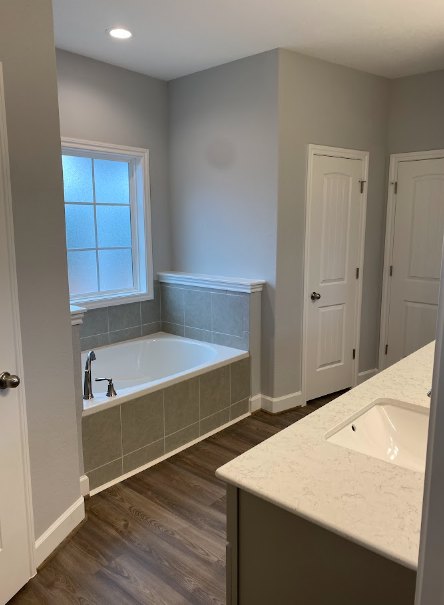 Bathroom with freestanding white bathtub, rectangular white sink on wood vanity, frosted glass window, wood flooring, white door with silver hardware, light tile walls