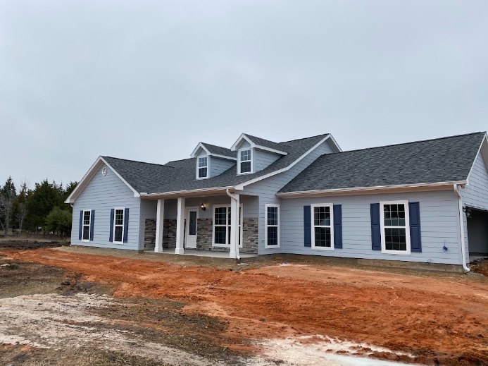 Partially built house with blue roof, white framed windows, blue shutters, exposed dirt yard, surrounded by trees