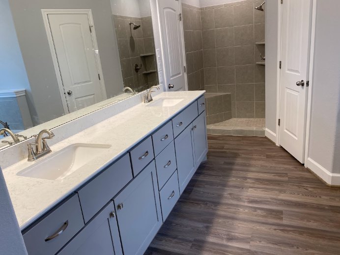 Bathroom with two undermount sinks set in a quartz countertop, modern chrome faucets, light wood cabinetry, glass-enclosed shower with tile walls, and neutral tile flooring