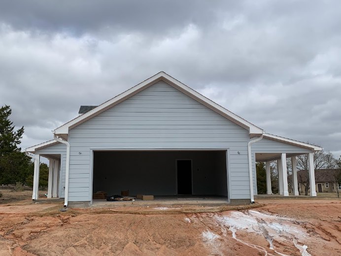Partially built house with exposed framing and garage, muddy ground in foreground, cloudy sky overhead, American Gothic House visible in background, trees and shed nearby.