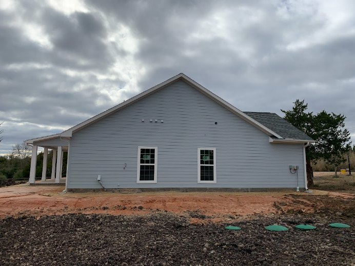 Grey house under construction with white trim and pillars, cloudy sky overhead, window visible on exterior wall, rooftop partially shown, tree and historic Rockingham Meeting House