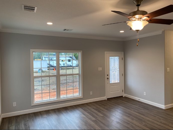 Neutral-toned room featuring a ceiling fan, large window with white trim, light-colored flooring, and simple wall molding.