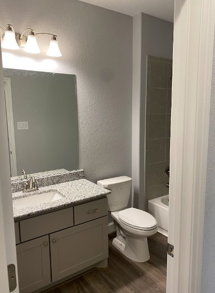 Modern bathroom with white toilet, rectangular sink on wood vanity, light gray tile floor, three-bulb wall sconce, white walls, and light switch.