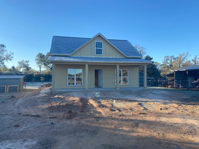 Partially built house with exposed wooden framing, shingled roof, covered porch, white-framed window, dirt yard with puddle, chain-link fence, and mature trees in the background
