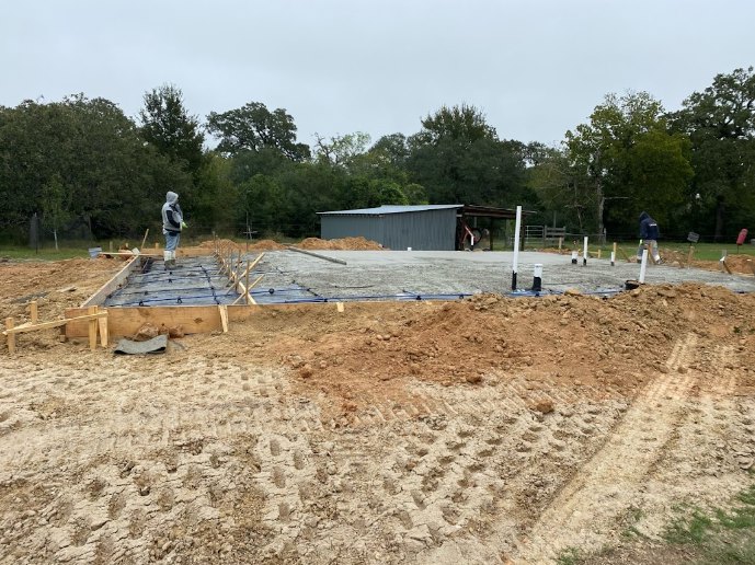 Man standing on muddy construction site in front of partially built custom home with exposed foundation, grass, and trees in background