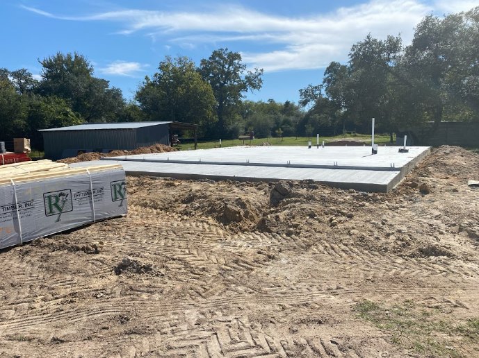Concrete foundation slab surrounded by dirt piles, grassy patches, and scattered construction materials; trees and blue sky in background.