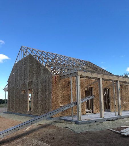 Partially built house with exposed wooden framing, exterior walls, ladder leaning against structure, cloudy sky overhead, construction materials scattered on ground