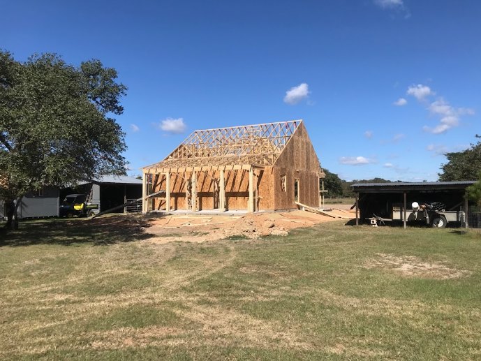 Wood-framed house under construction with exposed beams, partially built roof, green grass, dirt patches, leafy tree, and pickup truck parked nearby