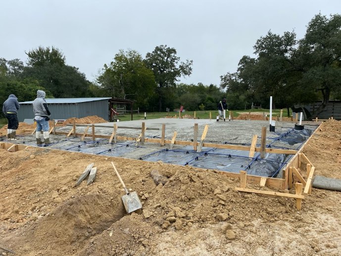 Men working on a home foundation surrounded by soil, construction materials, and trees under a clear sky