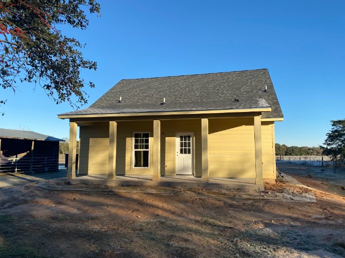 Yellow siding house with white trim, covered front porch, white entry door, metal fence, dirt patch in front yard, large tree with red berries in backyard, gable roof, multiple