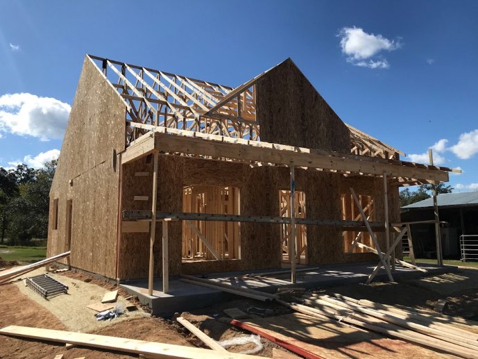 Wood-framed house under construction with exposed beams, pile of lumber on dirt ground, temporary fencing and utility pole in foreground, blue sky with scattered clouds overhead