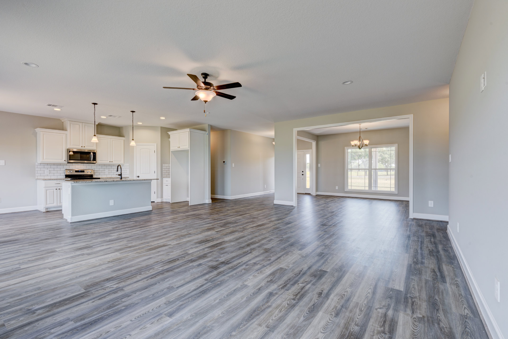 Spacious open floor plan featuring wood flooring, white walls, ceiling fan with light fixture, marble kitchen countertop, window with overhead light, and built-in microwave