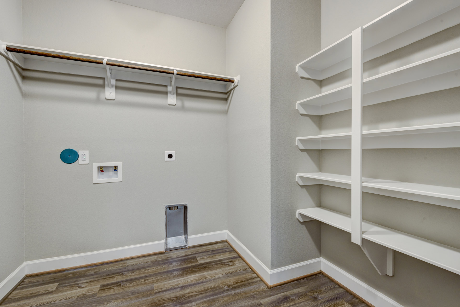 Built-in white shelving along a plaster wall, empty closet with multiple shelves, light wood flooring, recessed ceiling, and visible water valves in a white utility box.