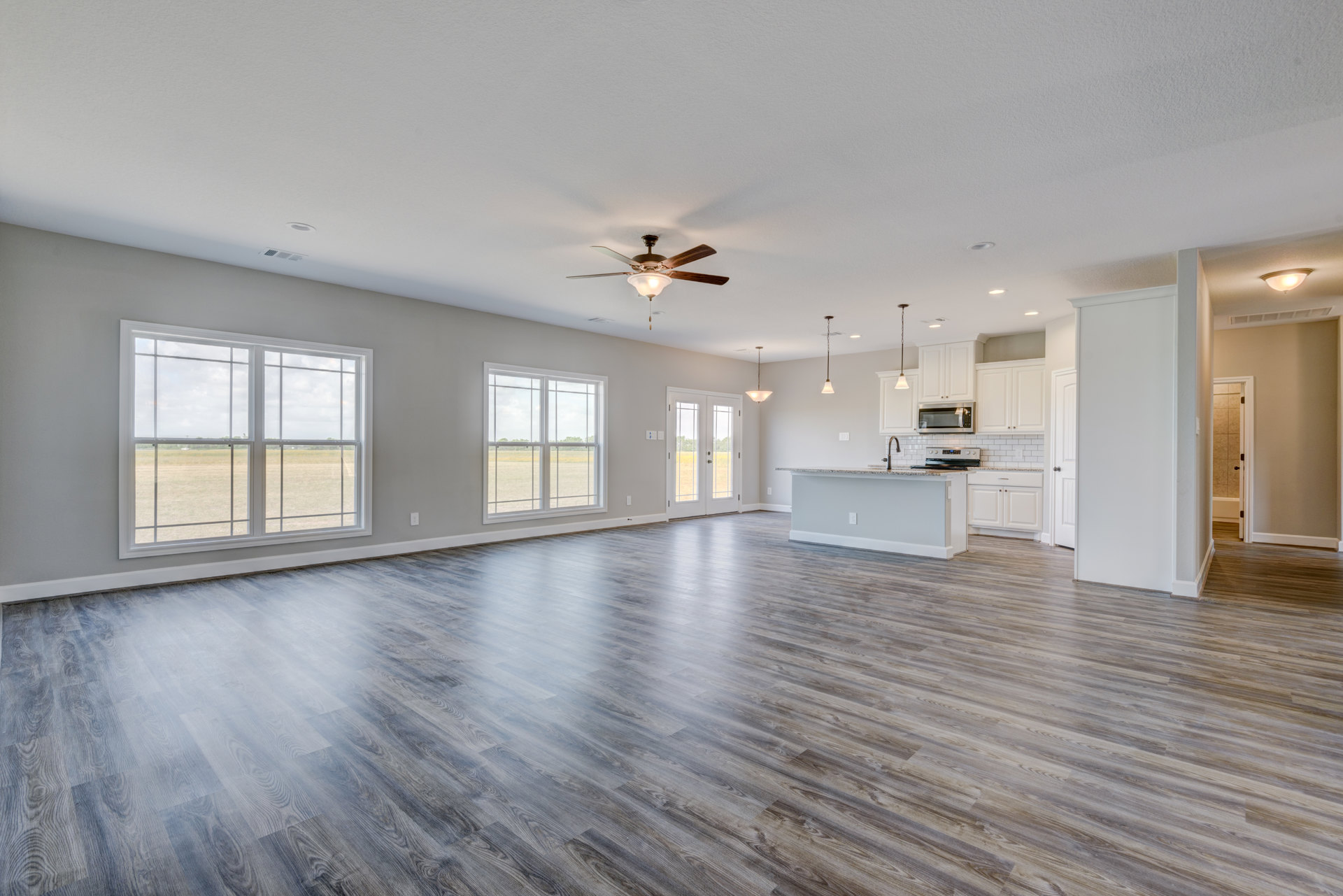 Spacious open-plan room featuring wood flooring, white plaster walls, large windows overlooking grassy field, modern kitchen with cabinetry, and ceiling fan with light fixture