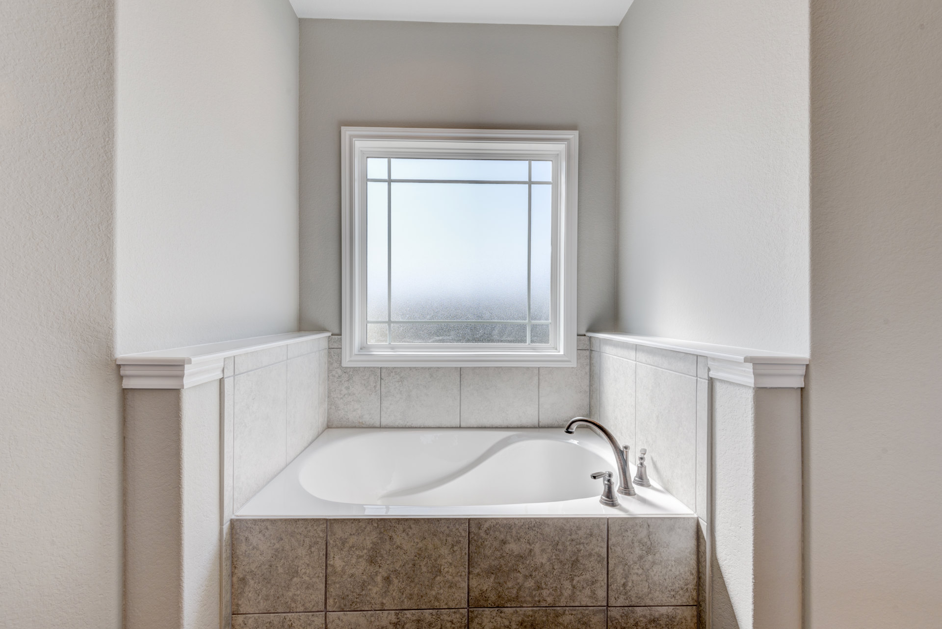White freestanding bathtub with chrome faucet on light tile floor, frosted glass window above, neutral wall finishes