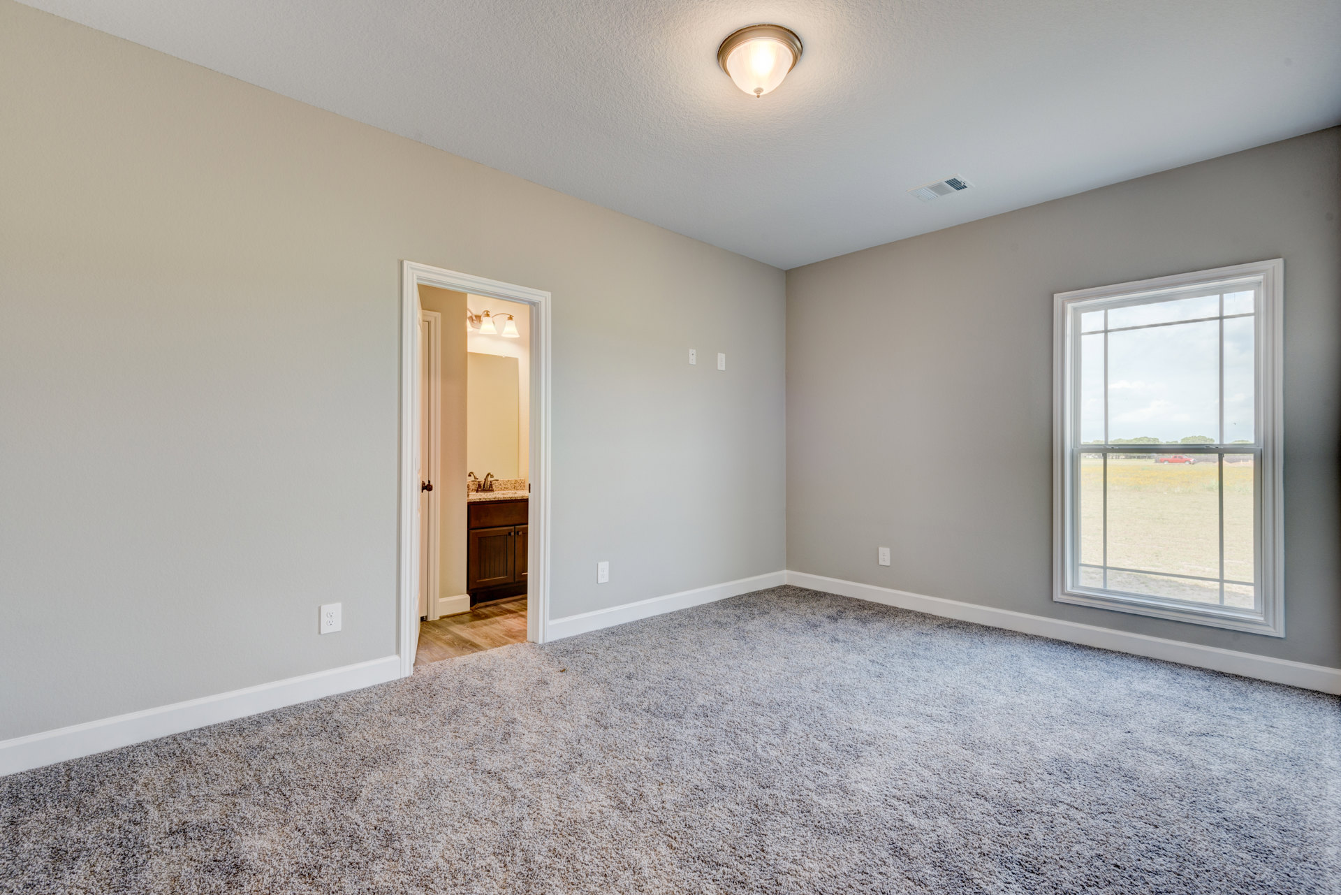 Carpeted room with white walls, paneled door, large window showing a red truck outside, ceiling-mounted light fixture, and built-in cabinet; bathroom with white sink and framed