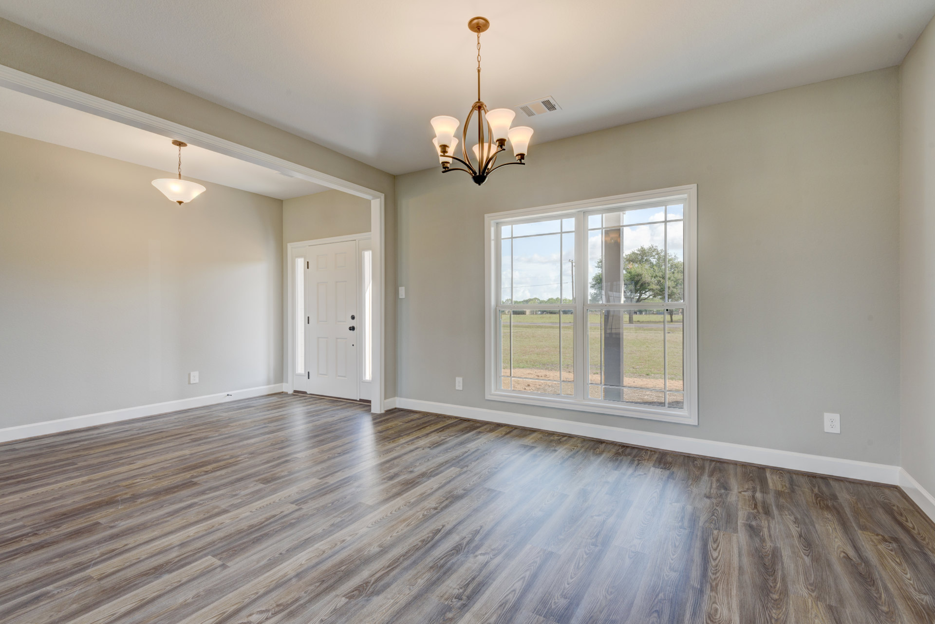 Wood flooring in a spacious room with white walls, decorative molding, a black-knobbed white door, large window overlooking a grassy field, and a modern chandelier hanging from a