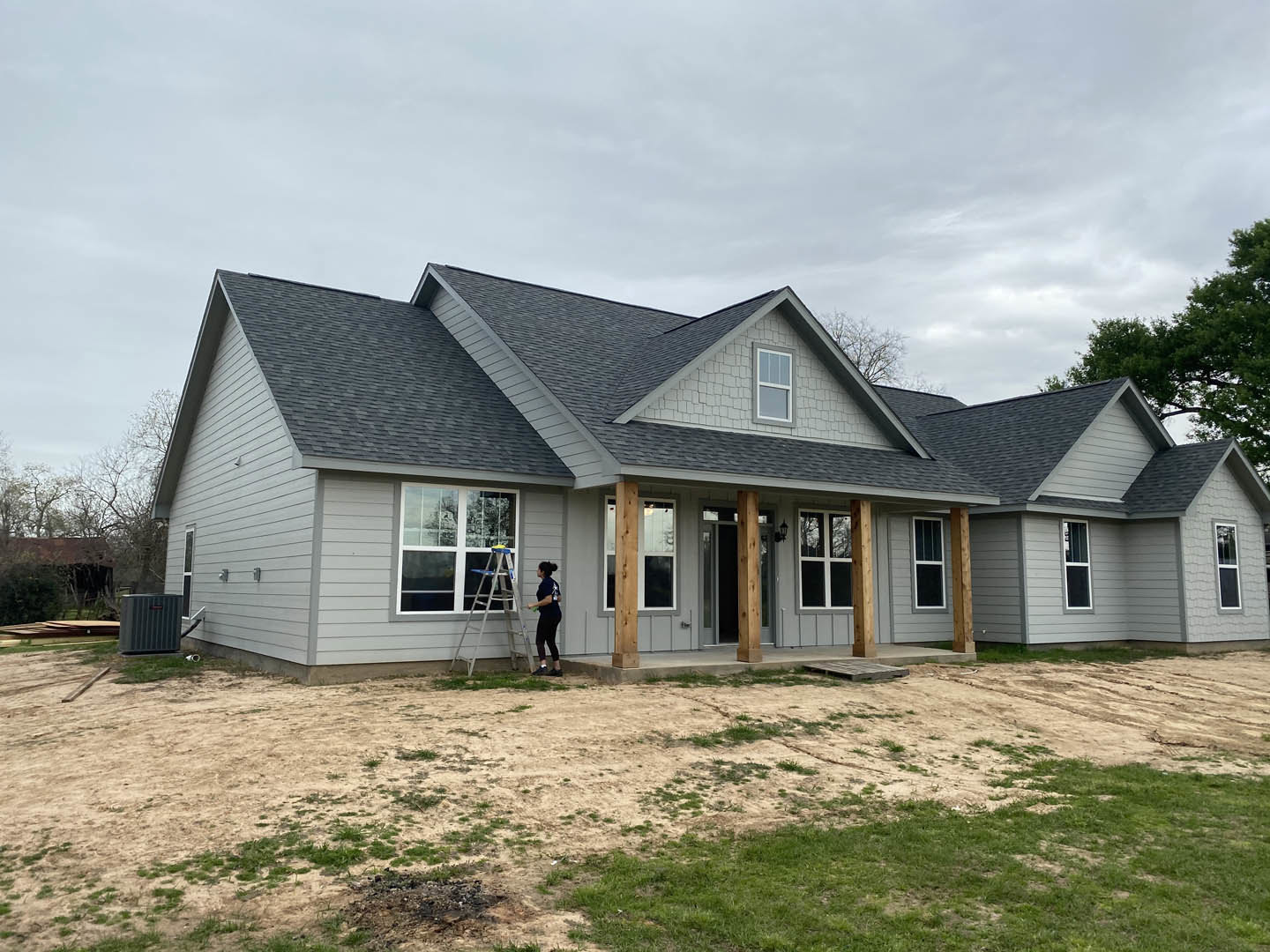 Woman on ladder working near window of white clapboard house, Robert Frost Farm visible in background, surrounded by trees and cloudy sky.
