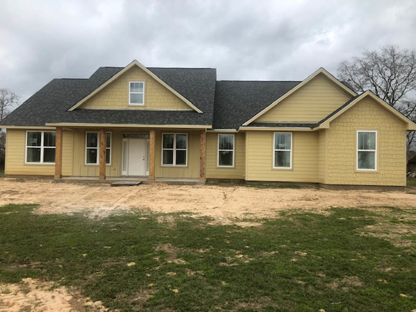 Partially built house with black shingle roof, white siding, white framed windows, and white door, surrounded by grass and dirt under a cloudy sky