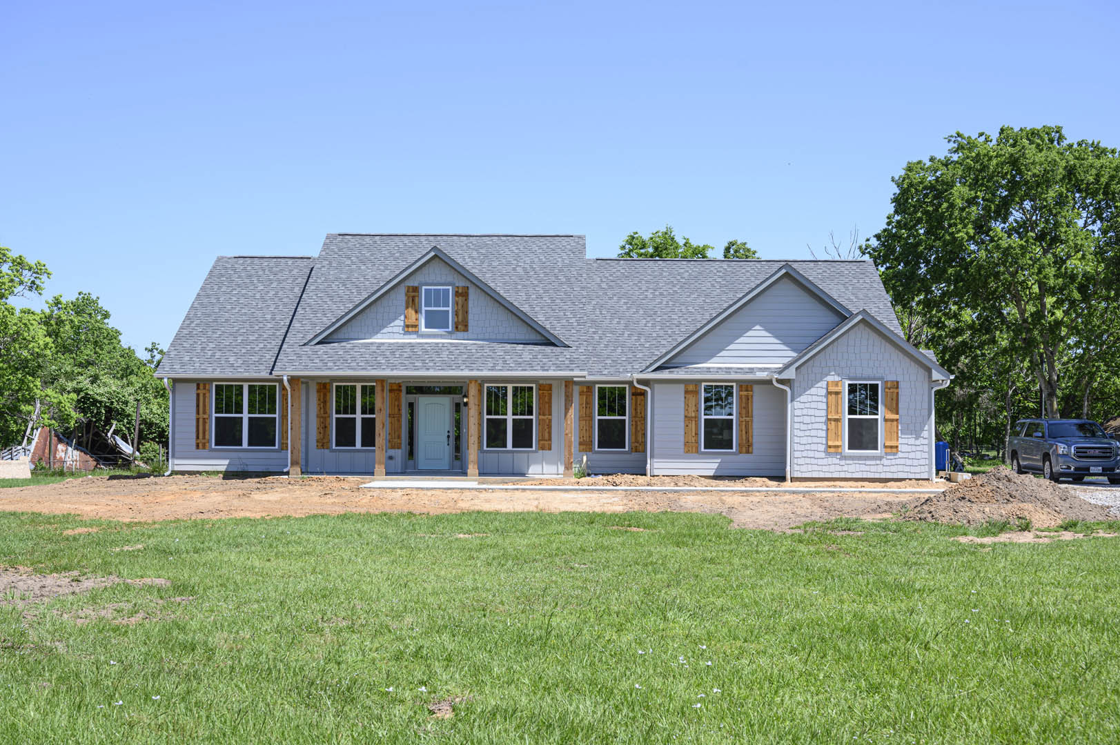 Two-story house with grey roof, white trim windows, covered porch, green lawn, mature tree, and silver SUV parked on dirt driveway